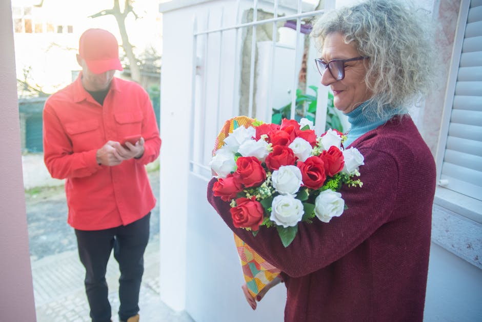 A woman with curly gray hair, glasses, and a red sweater holds a vibrant bouquet of fresh red and white roses wrapped in patterned tissue paper, standing outside near a white window frame. A man dress