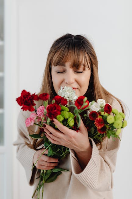 A woman with light brown hair and bangs, dressed in a beige sweater, holds a large bouquet of fresh flowers close to her face with her eyes closed and a gentle smile. The bouquet features a mix of vib