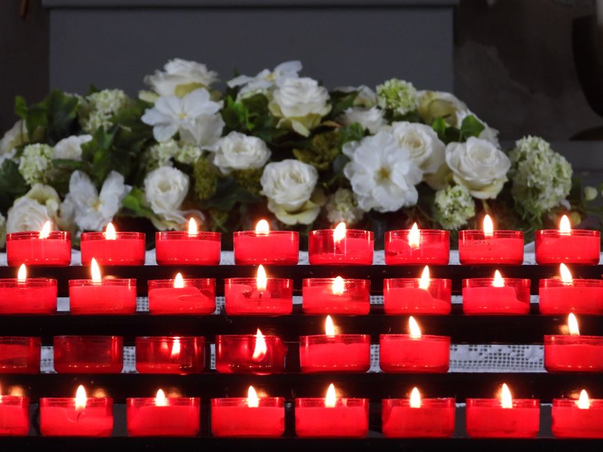 A floral arrangement of white roses, hydrangeas, and other delicate white flowers with lush green foliage, arranged on a flat surface behind a row of lit red votive candles. The flowers appear fresh,