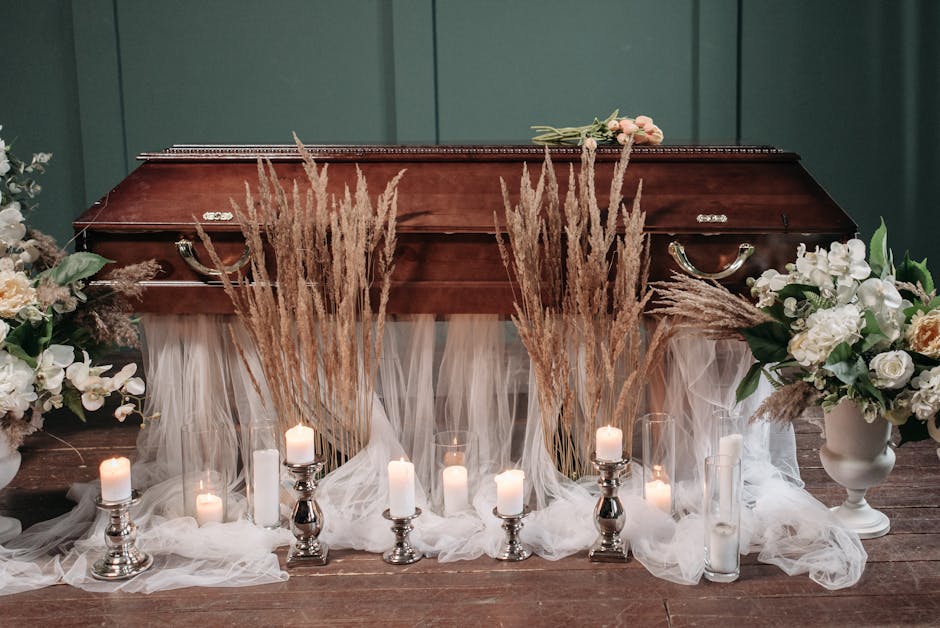 A funeral tribute arrangement featuring a wooden casket placed on a wooden floor, surrounded by white and cream floral bouquets with roses, carnations, and lilies, arranged in white ceramic vases. Tal
