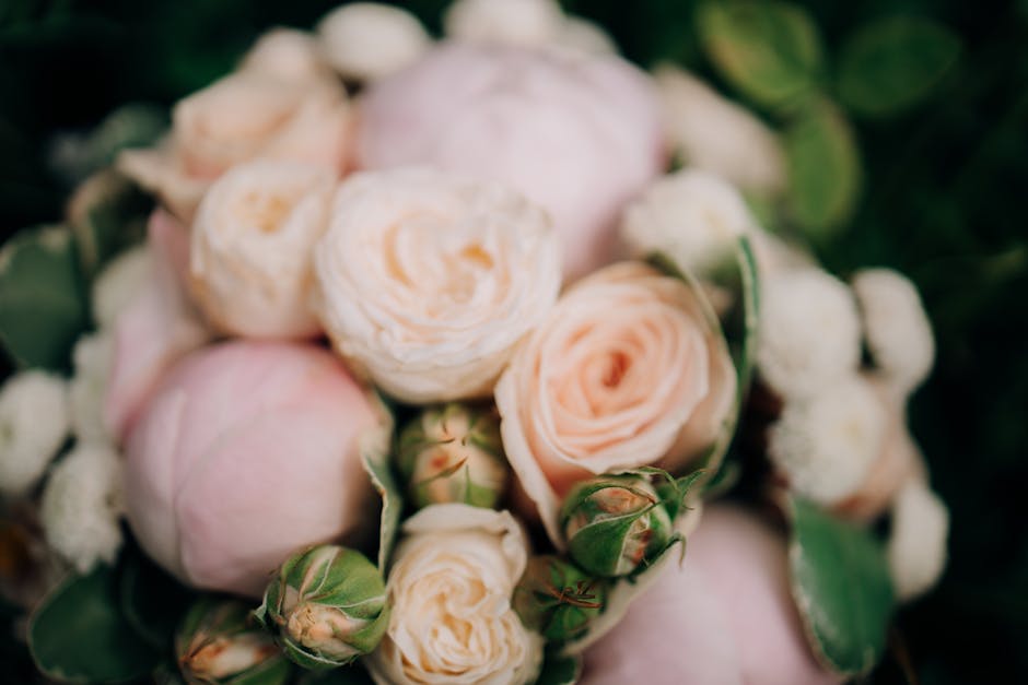 A close-up of a floral arrangement featuring soft pink and cream roses with tightly curled petals and a delicate texture, complemented by small white filler flowers and glossy green leaves. The bouque