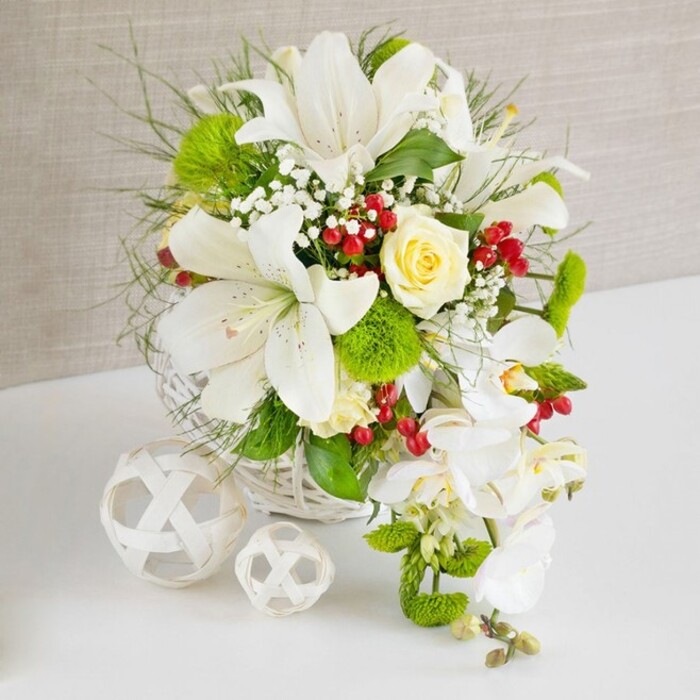 This image presents a lavish white and green floral composition arranged in a round white wicker basket, positioned on a smooth white surface with a softly textured beige backdrop, reminiscent of a well-lit North Woolwich reception room or riverside restaurant. At the heart of the design, large white lilies open fully, their trumpet-shaped petals stretching outward and slightly back, revealing speckled interiors and golden stamens that add a subtle depth of colour. Tucked between these lilies, a single creamy ivory rose blooms in perfect form, its petals spiralling gently from the centre and creating a warm focal point that contrasts softly with the crisp white around it. Flowing from the right side of the basket, an elegant cascade of white phalaenopsis orchids descends in a gentle curve, their broad, smooth petals and pale centres forming a soft, waterfall-like effect that gives the arrangement a romantic, bridal feel, ideal for a top table in North Woolwich. Surrounding and balancing these hero flowers, layers of greenery spill outward-fine, wispy grasses, feather-light fronds, and broader, darker leaves-creating a sense of abundance and natural movement. Bright green pom-pom blooms, reminiscent of dianthus or green trick, are nestled among the foliage, their tufted, cushion-like texture adding tactile interest and visual contrast. Sprays of tiny white gypsophila weave delicately through the bouquet, filling spaces and imparting an airy, cloud-like quality. Clusters of deep, ruby-red berries stand out against the greenery, bringing warmth and a slightly festive touch that suggests celebration and joy. To the left on the table, two decorative white wicker spheres rest side by side, one slightly larger than the other, their open lattice design echoing the basket and adding a sculptural element. Overall, the piece feels refined, tranquil, and meticulously composed, ideal as a wedding table arrangement or luxurious centrepiece for local events.